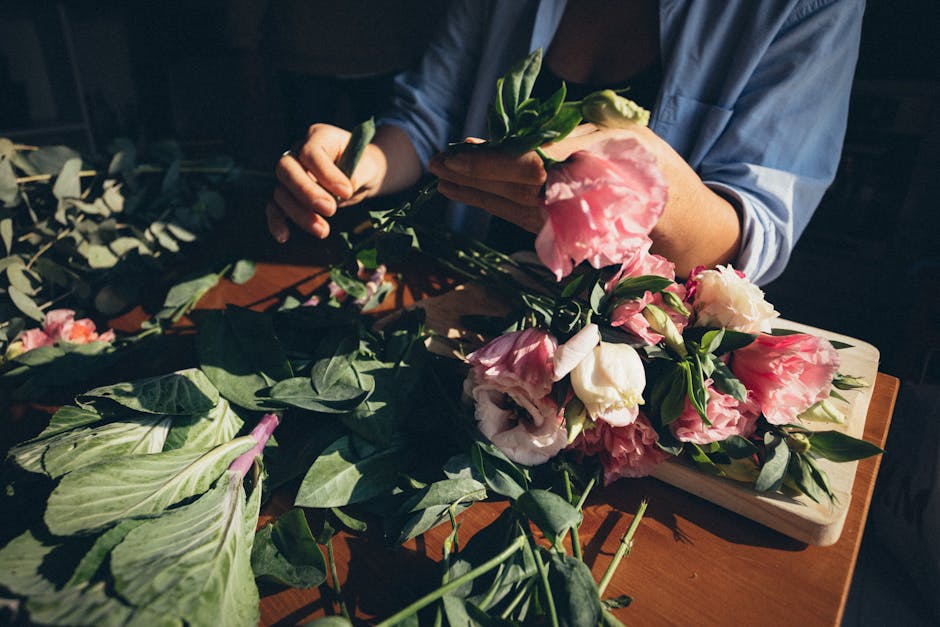 A person arranging a floral bouquet featuring large, ruffled pink and white lisianthus flowers with lush green foliage on a wooden surface. The arrangement surrounds a wooden board, with their hands g