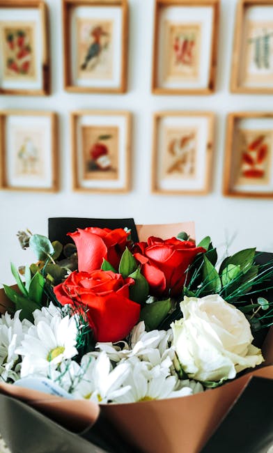 A close-up view of a floral bouquet arranged with vibrant red roses and white chrysanthemums, wrapped in soft peach-colored paper with green foliage accents. The bouquet is held in a person's hand, po