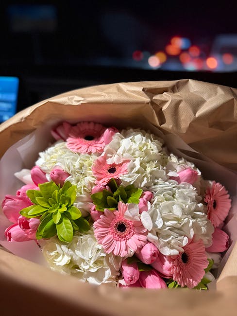 A floral bouquet arranged with white hydrangeas, pink gerbera daisies, and green foliage, wrapped in brown kraft paper with pink tissue paper peeking through. The flowers are fresh, with the petals an