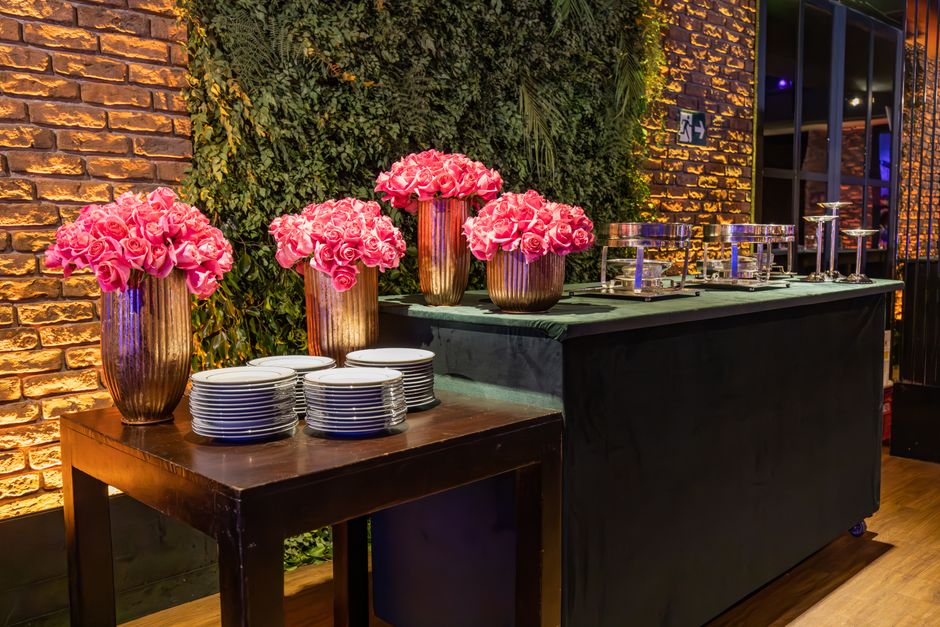 A floral arrangement featuring three tall, cylindrical, rose-golden vases filled with vibrant pink roses, placed on a polished wooden table alongside white stacked plates. To the right, a black table 