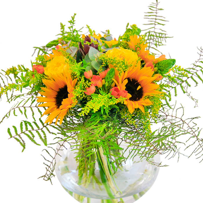 From the moment you set eyes on this sun-drenched bouquet, there's an undeniable sense of cheer and celebration-evocative of a perfect day in Bickley when sunlight dances over garden beds. Each sunflower, with its rich, chocolate-brown center and radiant yellow petals, seems to reach outward in joyful exuberance. Nestled alongside, soft yellow roses open in classic spirals, their layers of petals inviting the touch and aroma that recall the scent of local blooms caught on a gentle breeze down Bickley Road. Glossy red hypericum berries twinkle among the greens, adding contrast and a playful spark that calls to mind laughter-filled afternoons in local parks. The lush tapestry of foliage includes fronds of emerald fern and airy sprigs-possibly goldenrod-with frothy blossoms and slender green spikes, offering texture and dynamism to the overall shape. Silvery eucalyptus leaves provide a cool contrast, their subtle bluish tint tempering the warm tones. Everything is perfectly arranged in a clear, globe-shaped glass vase, the stems visible in crisp water and rooting the vivid arrangement in freshness and clarity. Crafted with the expert hands of Bickley Florist, this centerpiece is ideal for those moments-birthdays, anniversaries, or spontaneous celebrations-when joy and togetherness deserve to be at the heart of the moment in Bickley homes.